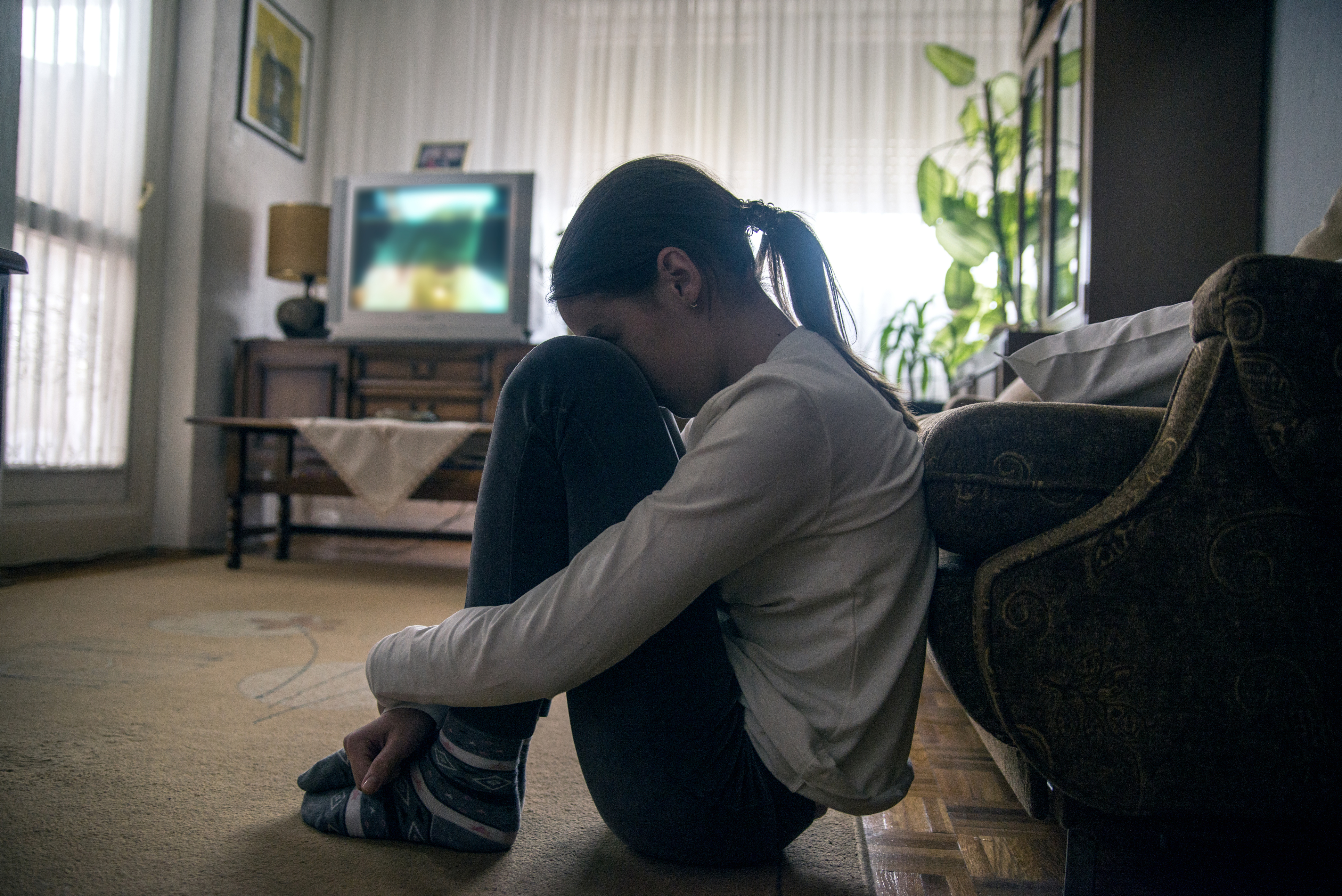 A young person sitting against a sofa with their head on their knees